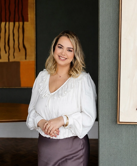 Foto da Ana Carolina sorrindo, de blusa branca e saia de cetim marro, em uma sala com um grande quadro ao fundo