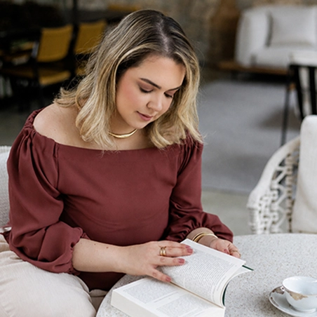 Foto da Ana Carolina sentada em uma cadeira com um livro apoiado na mesa, lendo. Ao lado do livro tem uma xícara de chá